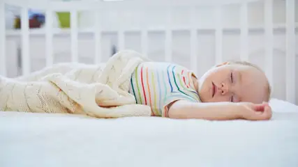 Baby mit gestreiftem Body schläft auf dem Rücken in einem weißen Gitterbett und ist mit einer hellen Strickdecke zugedeckt.