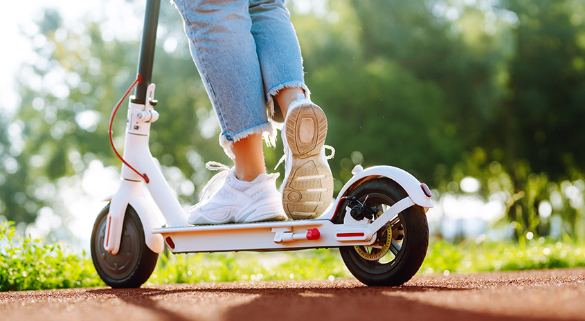 Ein weißer E-Scooter und darauf zu sehen 2 Beine bis zu den Knien sichtbar mit weißen Turnschuhen und einer hellblauen Jeans, dahinter sieht man unscharf grüne Bäume