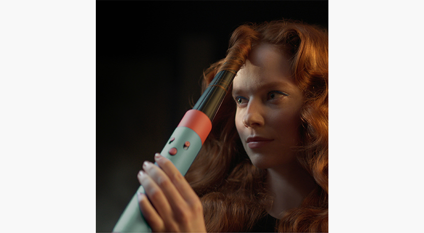 Eine Frau benutzt ein blaugrünes und orangefarbenes Lockenstab, um ihr Haar zu locken. Der Hintergrund ist dunkel.