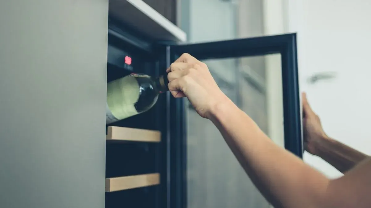 Hände legen eine helle Weinflasche in einen geöffneten Weinkühlschrank mit Holzregalen und roter Anzeige.