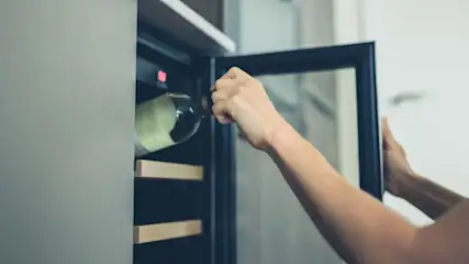 Hände legen eine helle Weinflasche in einen geöffneten Weinkühlschrank mit Holzregalen und roter Anzeige.