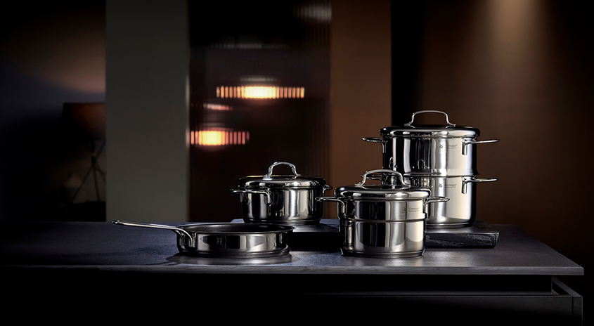 Auf einer dunklen Oberfläche stehen vier silberne Töpfe unterschiedlicher Größe, darunter eine Bratpfanne ohne Deckel.