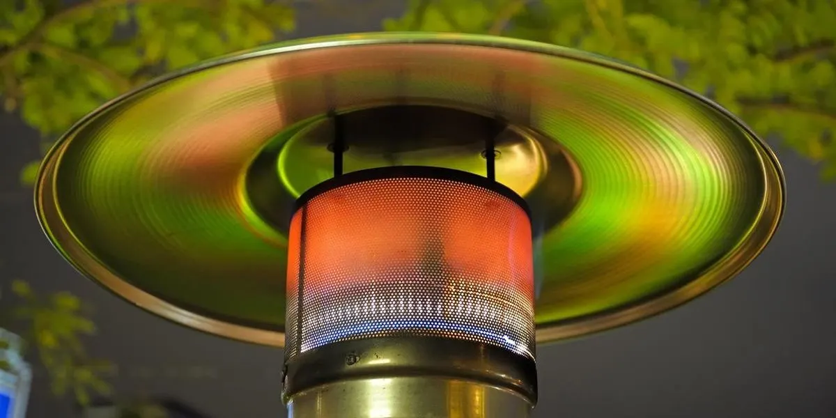 Ein Heizstrahler mit orange-blauem Licht und einer Metallplatte, die grün und gelb reflektiert. Grüne Blätter im Hintergrund.