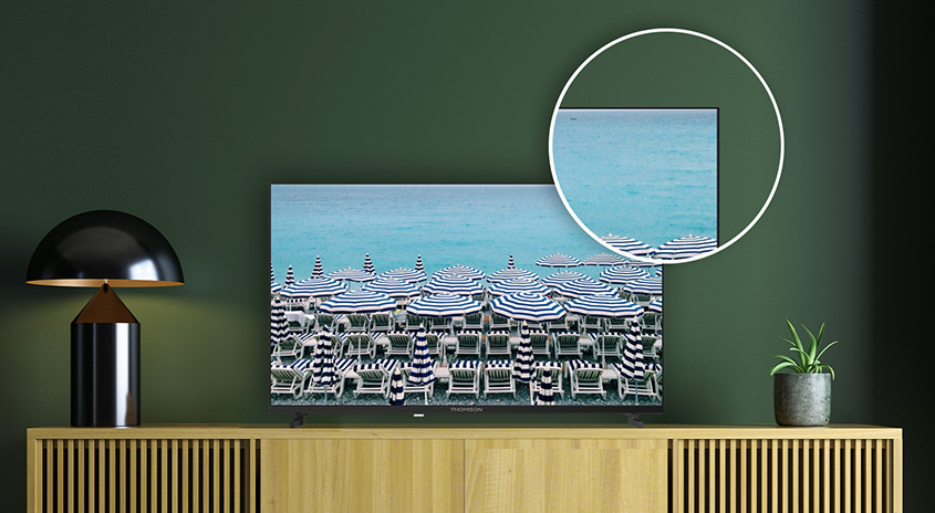 Auf einem Holzschrank steht ein Fernseher, der einen Strand mit blau-weiß gestreiften Sonnenschirmen zeigt. Daneben stehen eine Lampe und eine Zimmerpflanze.