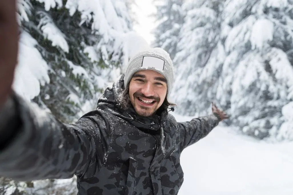 Ein lächelnder Mann im grauen Beanie und Tarnjacke macht ein Selfie in einem verschneiten Waldweg mit schneebedeckten Bäumen.
