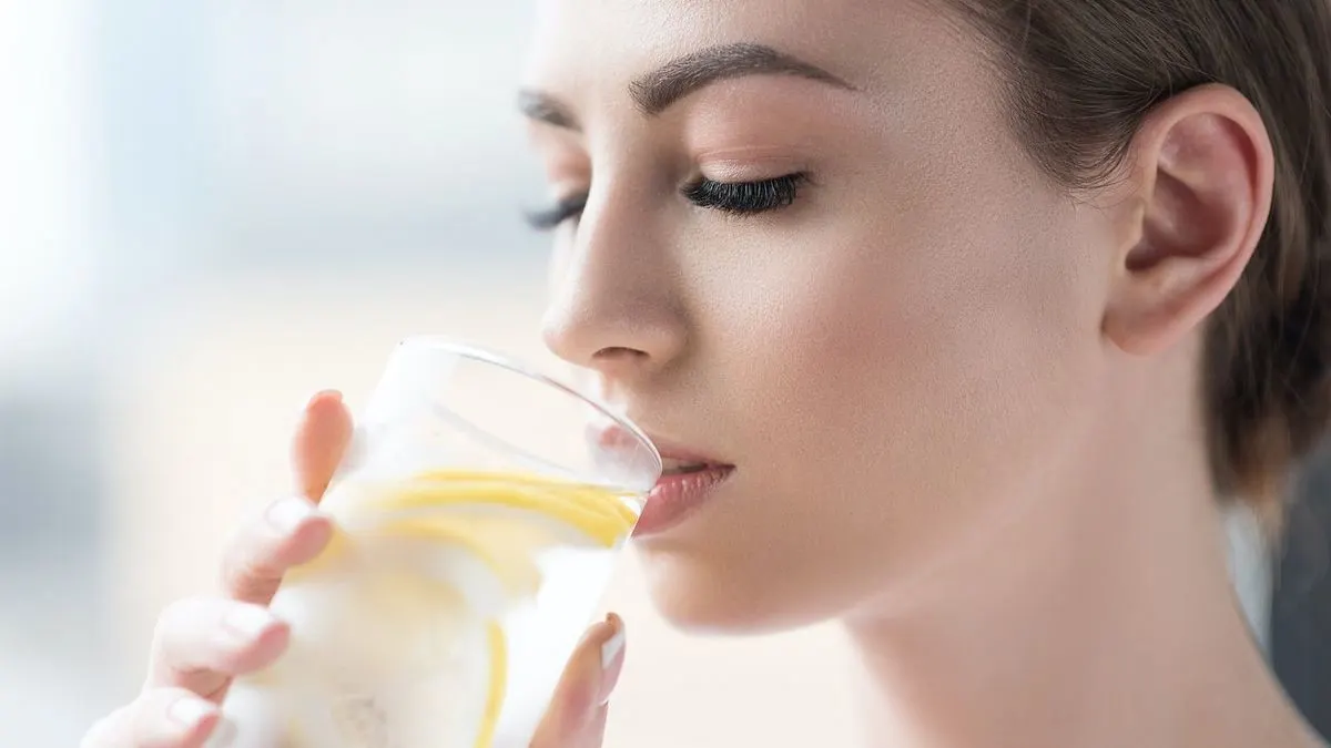 Person hält ein Glas Wasser mit Zitronenscheiben und Eiswürfeln in der Hand und führt es zum Mund.