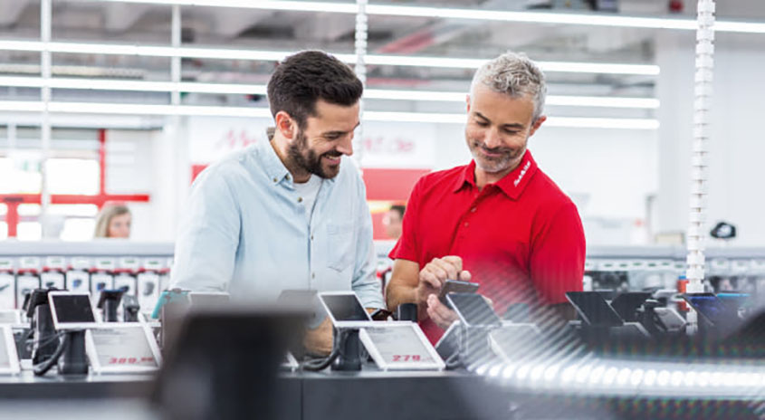 Ein Mann im hellblauen Hemd und ein Mann im roten Hemd betrachten elektronische Geräte in einem Geschäft.