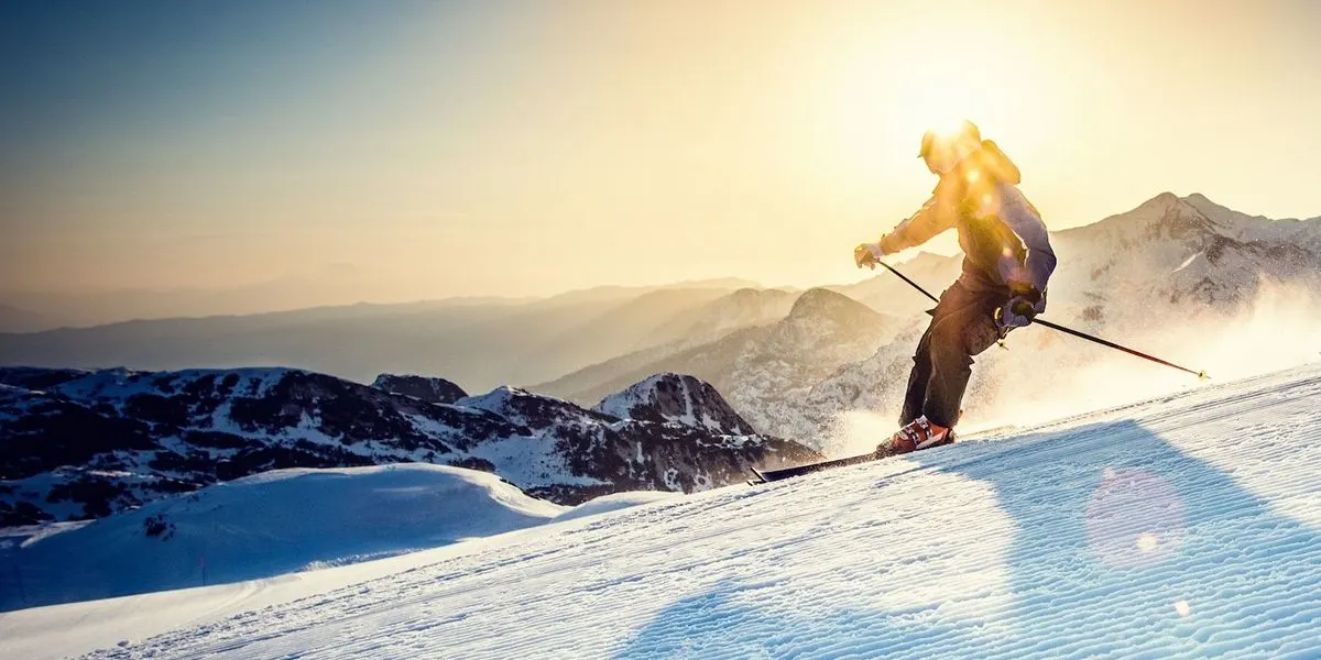 Person fährt bei Sonnenschein mit Skiern einen verschneiten Hang hinunter, im Hintergrund sind Berge zu sehen.