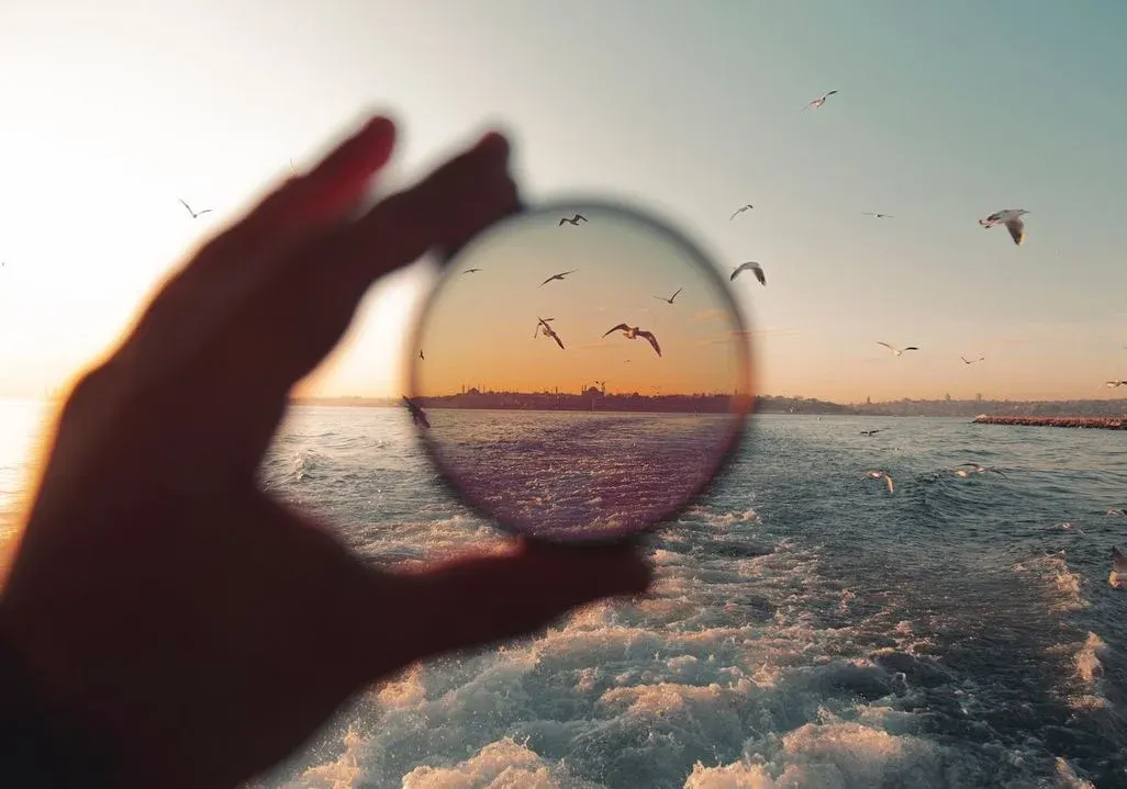 Hand hält runden Filter. Durch ihn: orangefarbener Sonnenuntergang, Stadt am Meer, fliegende Möwen. Außen: blauer Himmel, schäumendes Meer, Möwen.