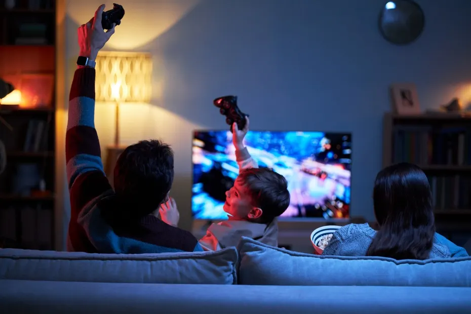 Drei Personen zocken abends auf dem Sofa; Controller hoch, TV zeigt Rennspiel.