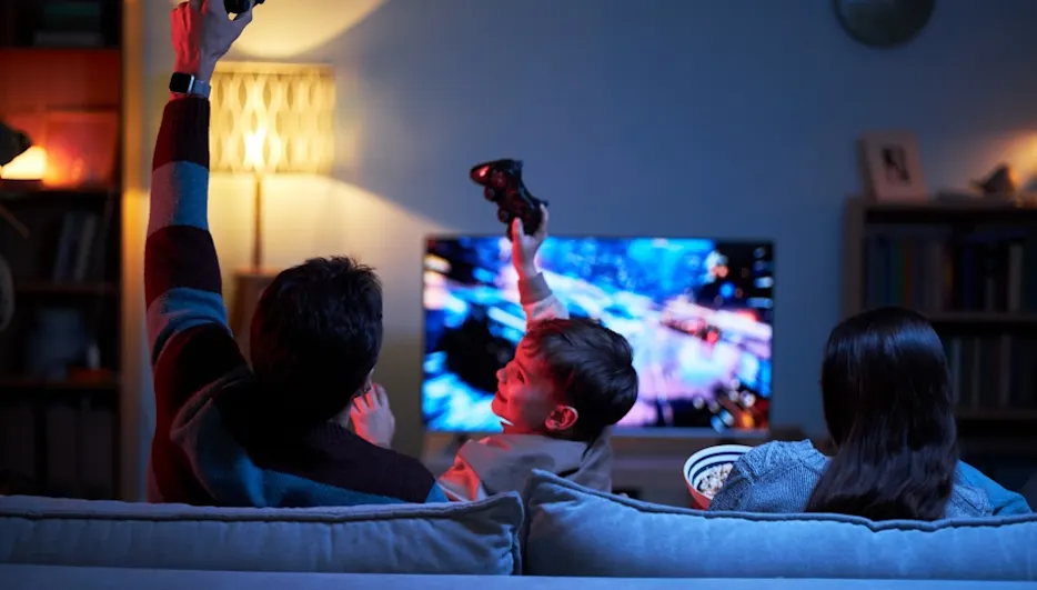 Drei Personen zocken abends auf dem Sofa; Controller hoch, TV zeigt Rennspiel.