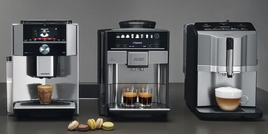 Drei silber-schwarze Siemens Kaffeemaschinen mit drei Kaffeespezialitäten in Gläsern und fünf bunten Macarons auf grauem Hintergrund.