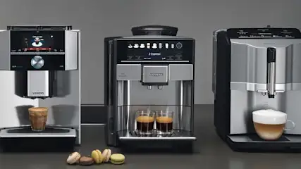 Drei silber-schwarze Siemens Kaffeemaschinen mit drei Kaffeespezialitäten in Gläsern und fünf bunten Macarons auf grauem Hintergrund.