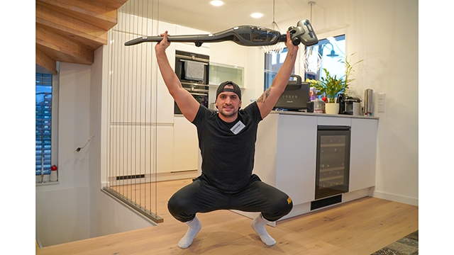 Ein Mann in Sportkleidung hockt und hält einen Gorenje Stiel-Staubsauger über seinen Kopf wie eine Langhantel.