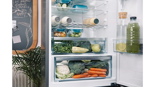 Ein offener Kühlschrank zeigt Regale mit Getränken, Gemüse und Salaten. Daneben steht eine Tafel mit einer Pflanze.