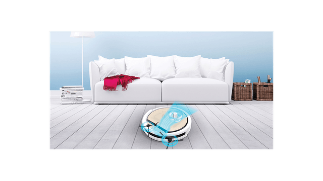 Ein Roboter-Staubsauger reinigt einen weißen Holzfußboden vor einem weißen Sofa mit Kissen und einer roten Decke.