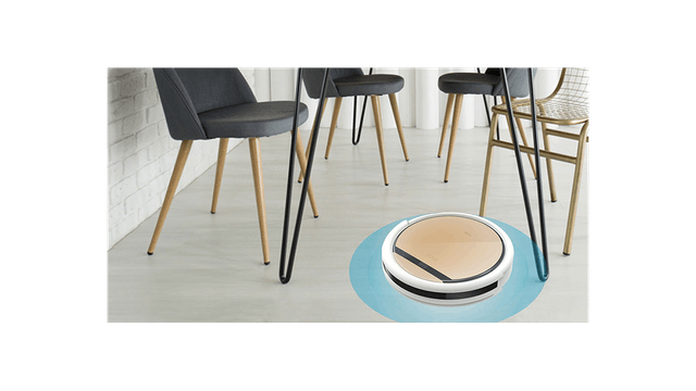 Ein runder Saugroboter auf einem hellen Boden unter Stühlen mit Holzbeinen. Eine weiße Ziegelwand ist im Hintergrund sichtbar.