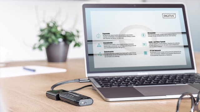 Ein Laptop zeigt eine Webseite neben einem USB-Gerät auf einem Tisch. Eine Pflanze und Dokumente sind im Hintergrund.