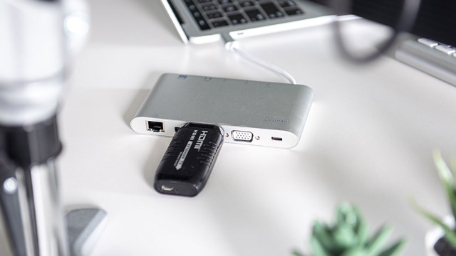 Auf einem weißen Tisch liegt ein silberfarbener Laptop-Hub mit einem angeschlossenen schwarzen HDMI-Adapter. Ein Laptop ist teilweise sichtbar.