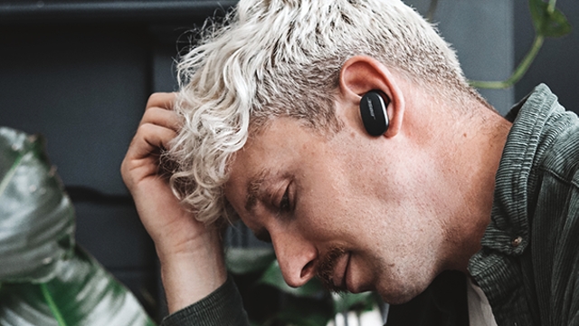 Ein Mann trägt einen schwarzen Ohrhörer und eine grüne Jacke. Er hält eine Hand am Kopf, mit Zimmerpflanze im Hintergrund.