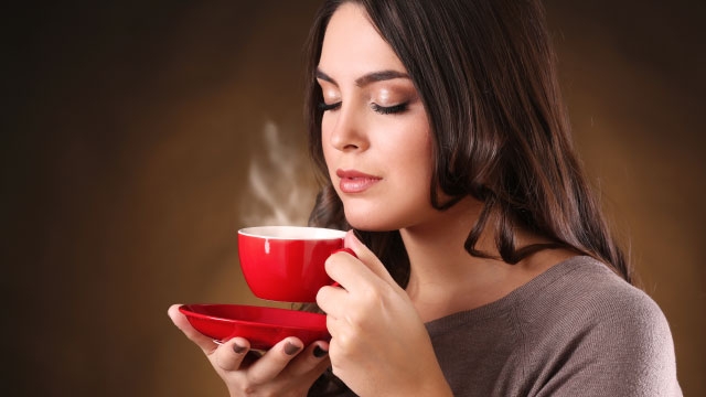 Eine Frau hält eine rote Tasse mit Untertasse. Dampf steigt auf, vor einem braunen Hintergrund.