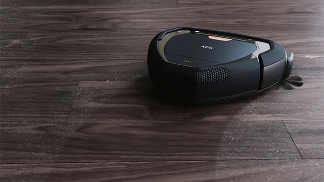 Ein schwarzer Staubsaugerroboter steht auf einem dunklen Holzboden mit horizontalen Brettern und leichten Glanzstellen.