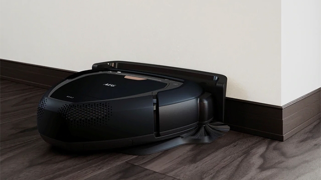 Ein schwarzer Saugroboter von AEG steht auf einem Holzboden vor einer weißen Wand mit dunkler Fußleiste.