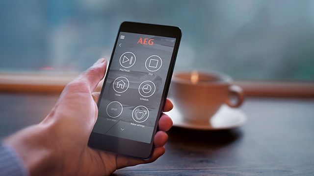 Eine Hand hält ein Smartphone mit einer AEG-App. Im Hintergrund ist eine Tasse auf einem Tisch zu sehen.