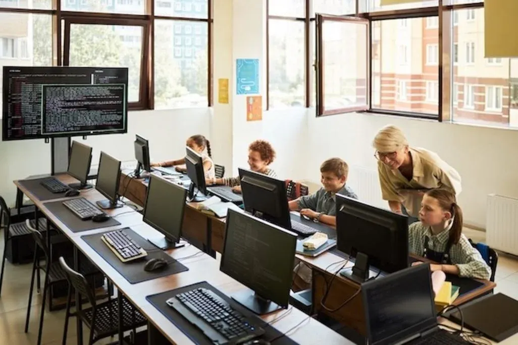 Volksschulkinder arbeiten an Desktop‑Computern im Computerraum, Lehrerin steht daneben und hilft einem Schüler.