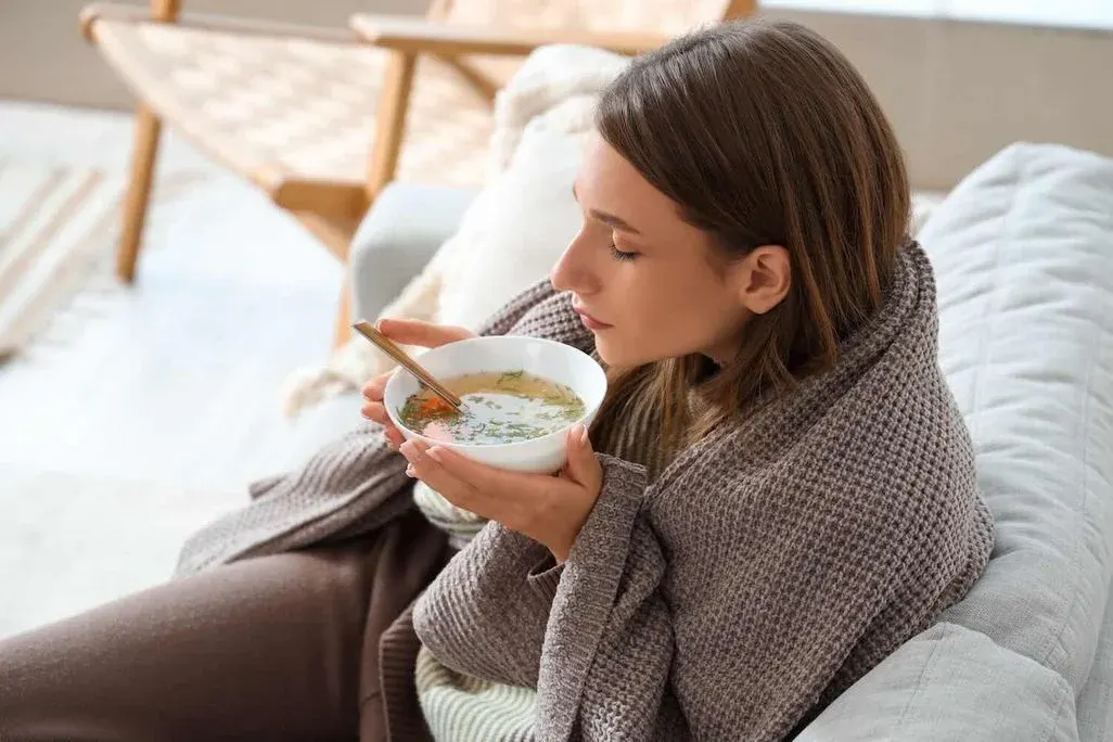 In eine Decke gehüllte Frau sitzt auf dem Sofa und isst eine warme Suppe aus einer Schüssel.