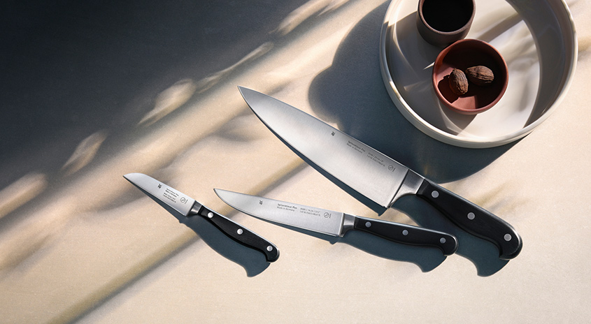 Drei Messer mit schwarzen Griffen liegen auf einer hellen Oberfläche. Eine Schale mit Nüssen steht neben einer Tasse auf einem Teller.