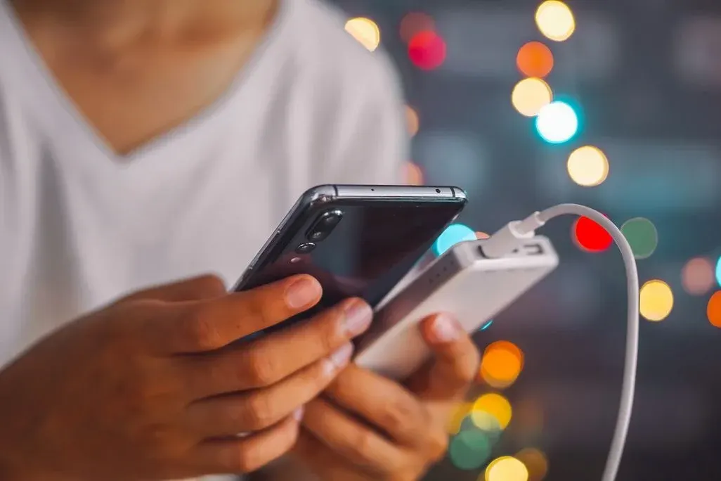 Person hält ein Smartphone in der einen Hand und eine angeschlossene Powerbank in der anderen, im Hintergrund bunte, unscharfe Lichter.