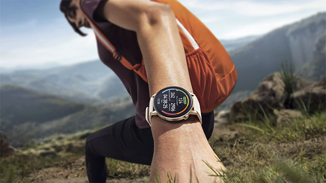 Eine Person mit Rucksack und Smartwatch wandert in den Bergen. Die Smartwatch zeigt Gesundheitsdaten an.