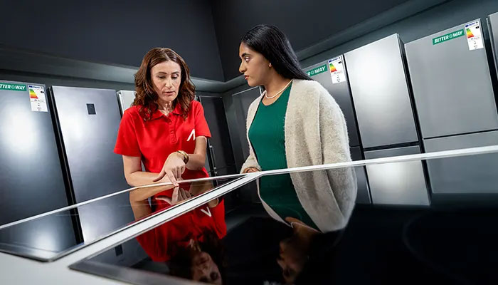 Zwei Frauen schauen sich in einem Geschäft Kochfelder an. Im Hintergrund sind Kühlschränke mit Energieetiketten zu sehen.