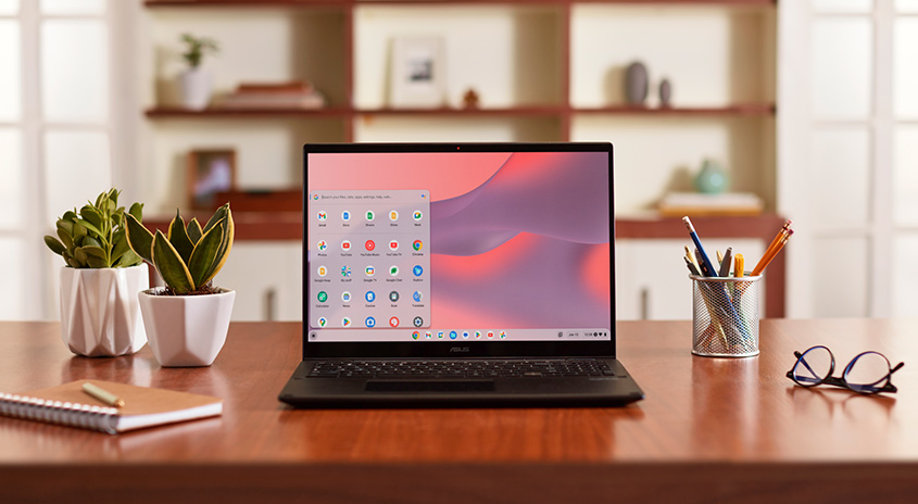 Ein Laptop steht auf einem Holztisch, flankiert von Pflanzen, einem Notizbuch, Stiften und einer Brille. Im Hintergrund befinden sich Regale.