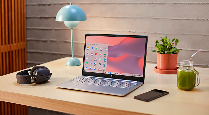 Ein Tisch mit Laptop, Kopfhörern, Lampe, Pflanze, Getränk und Handy vor einer Wand.