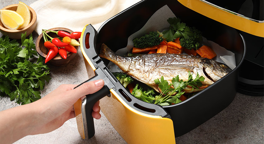  Hand zieht Korb einer gelben Heißluftfritteuse heraus, darin gebackener ganzer Fisch mit Gemüse und frischen Kräutern.