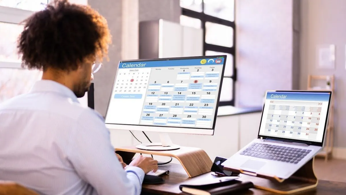 Ein Mann arbeitet mit Kalender-Apps auf einem Monitor und Laptop im Dual-Screen Setup für die Terminplanung.