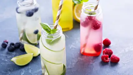 Vier Flaschen Infused Water: Blaubeer-Limette, Gurke-Basilikum, Himbeere und Zitrone. Früchte und Limettenspalten liegen auf dunkler Oberfläche.