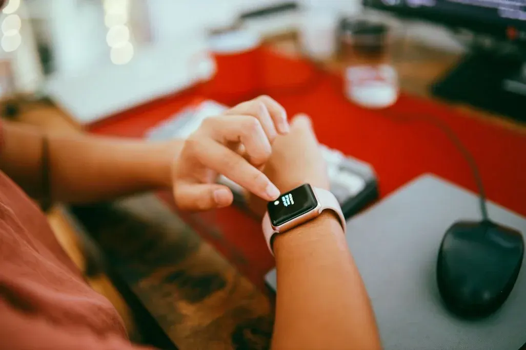 Person tippt mit dem Finger auf eine Smartwatch am Handgelenk, im Hintergrund ein Schreibtisch mit Computermaus.