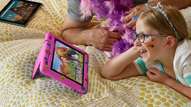 Ein Kind mit Brille schaut auf ein rosa Tablet mit Dinosaurierspiel. Ein anderes Tablet liegt im Hintergrund auf der gelb-weiß gemusterten Decke.