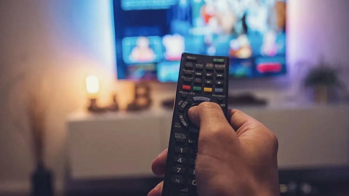 Hand haelt eine Fernbedienung vor eingeschaltetem Fernseher
