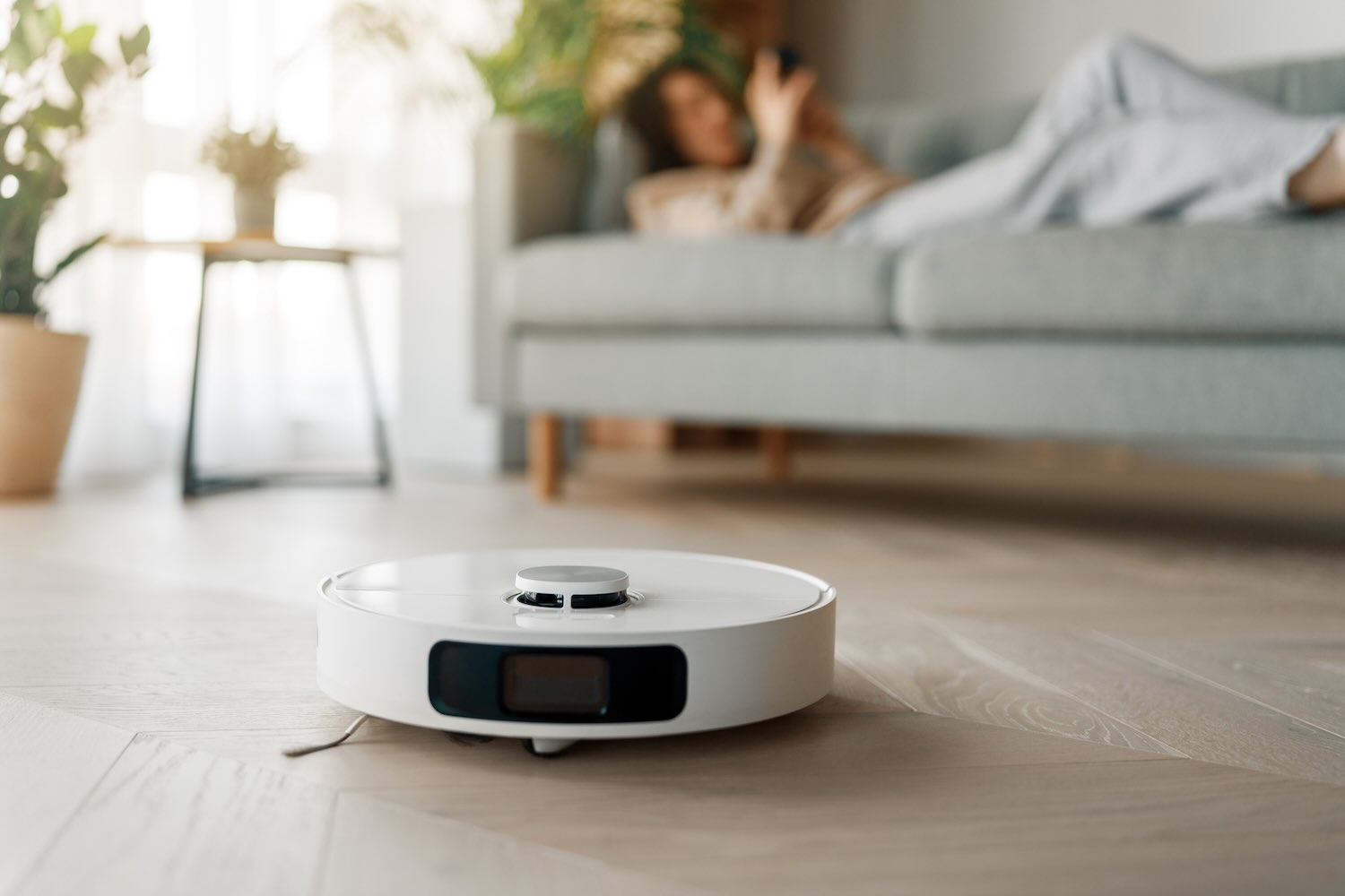 Ein weißer Saug-Wisch-Roboter fährt über einen Holzboden, im Hintergrund liegt eine Frau auf dem Sofa.