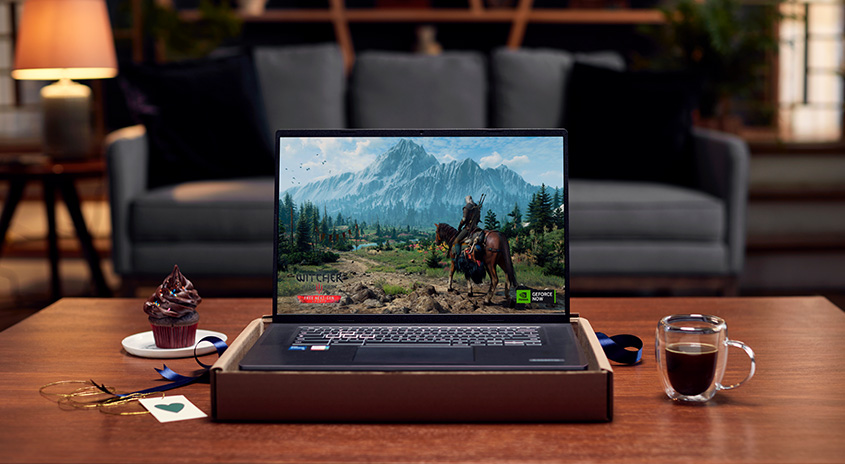 Ein Laptop zeigt ein Videospiel mit Bergen und einer Person auf einem Pferd. Auf dem Tisch stehen ein Muffin und eine Tasse Kaffee.