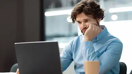 Ein nachdenklicher Mann stützt seinen Kopf in die Hand vor einem Laptop im Büro.