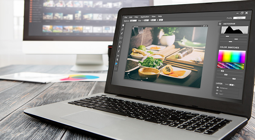 Ein Laptop zeigt ein Bildbearbeitungsprogramm mit einer Essensszene. Im Hintergrund ist ein Monitor auf einem Holztisch.