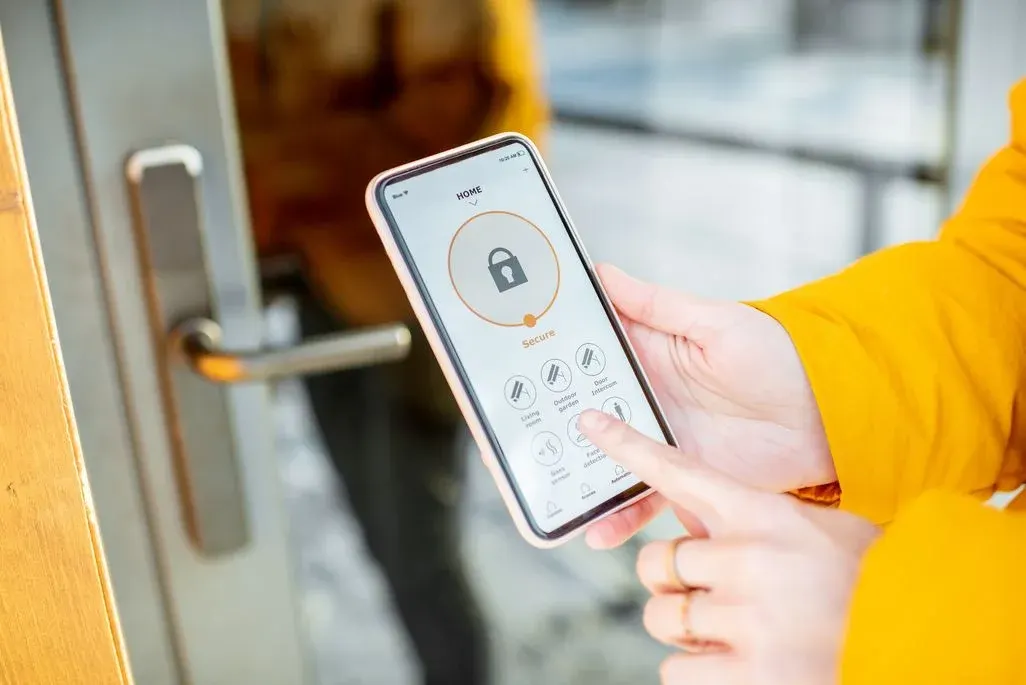 Hände halten ein Smartphone mit einer Smart-Home-App, die ein Schloss-Symbol und Steuerelemente anzeigt. Eine gelbe Jacke und eine Tür mit Griff sind sichtbar.