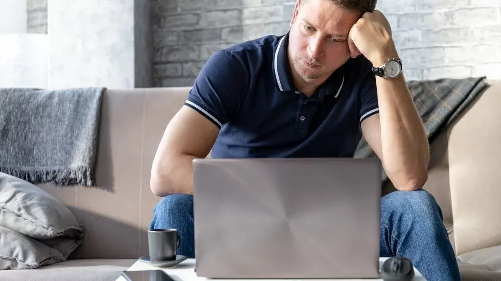 Mann in blauem Poloshirt und Jeans sitzt auf Sofa, stützt Kopf ab, blickt auf silbernen Laptop. Armbanduhr, Tasse, Maus auf Tisch.