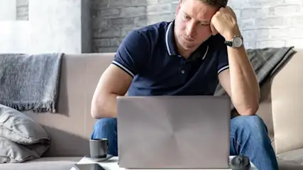 Mann in blauem Poloshirt und Jeans sitzt auf Sofa, stützt Kopf ab, blickt auf silbernen Laptop. Armbanduhr, Tasse, Maus auf Tisch.
