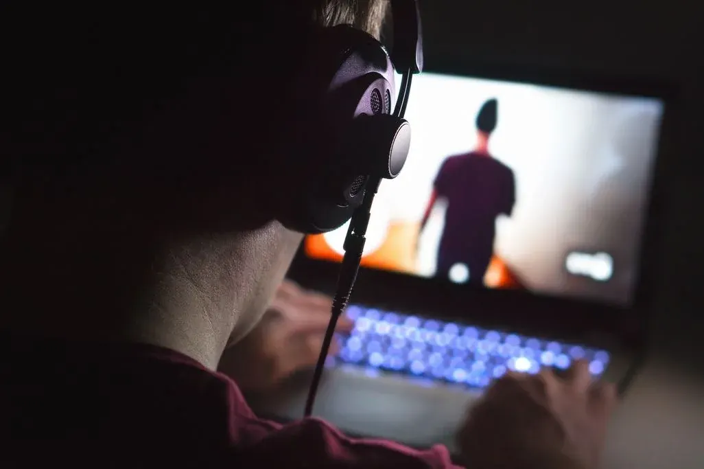 Person mit großen Kopfhörern sitzt in einem dunklen Raum vor einem aufgeklappten Laptop und spielt ein Videospiel; der Bildschirm zeigt eine unscharfe, stehende Figur, die Tastatur leuchtet.